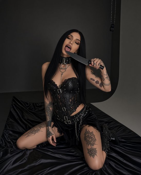 A woman with long dark hair wearing a black corset and skirt, posing with a knife near her face in a dark studio setting, with visible tattoos on her arms and legs.