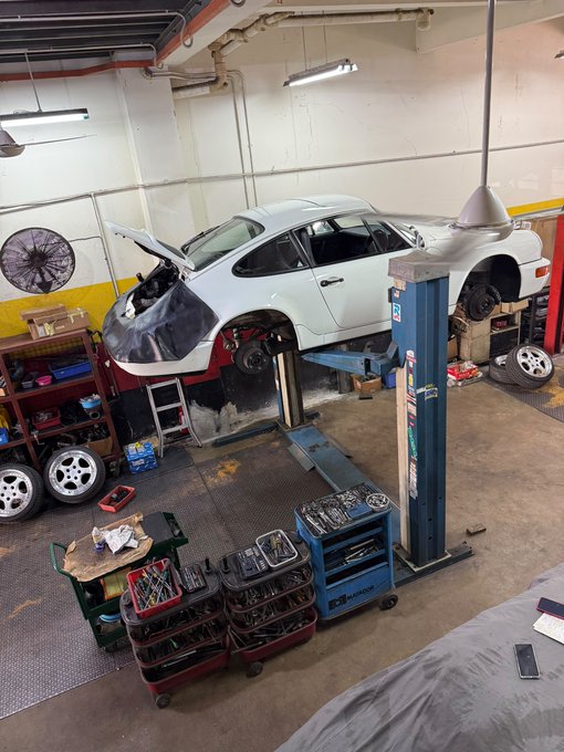 A white sports car is raised on a blue hydraulic lift inside a garage, surrounded by tool carts, scattered parts, and a wooden workbench.
