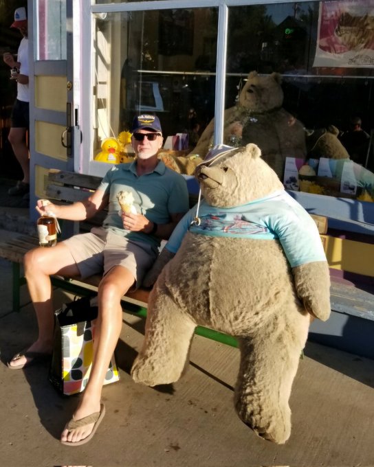 A man wearing an "Old Grand Dad" cap and "Let me have a Bourbon" shirt sits on a bench next to a large teddy bear, holding ice cream and a bottle of bourbon. The image humorously ties to the post's bullish Bitcoin celebration, as "Old Grand Dad" bourbon and the bear mascot visually represent the crypto trader's optimism about BTC holding above $80k and heading into a bull run.