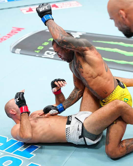 UFC fighter Joanderson Brito, wearing yellow and black shorts, throws a downward elbow strike against a bald opponent on the mat, with a referee observing the action amid Monster Energy and UFC branding on the canvas.