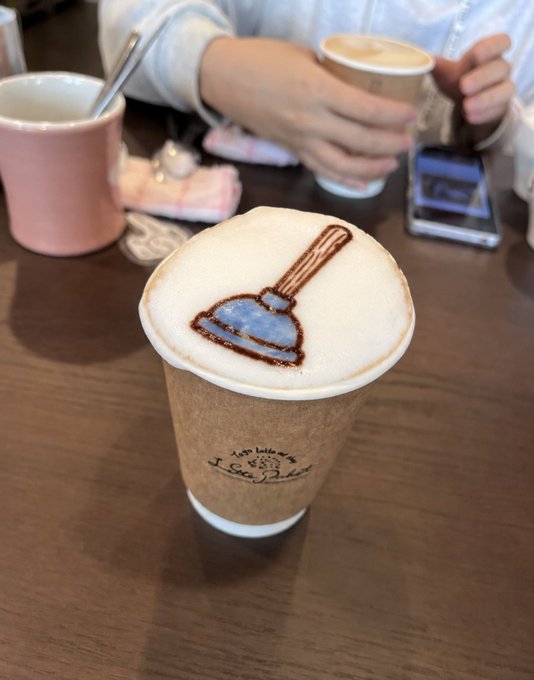A close-up of a latte with foam art depicting a broom, served in a paper cup at a café table, with another person's hands holding a similar cup in the background.
