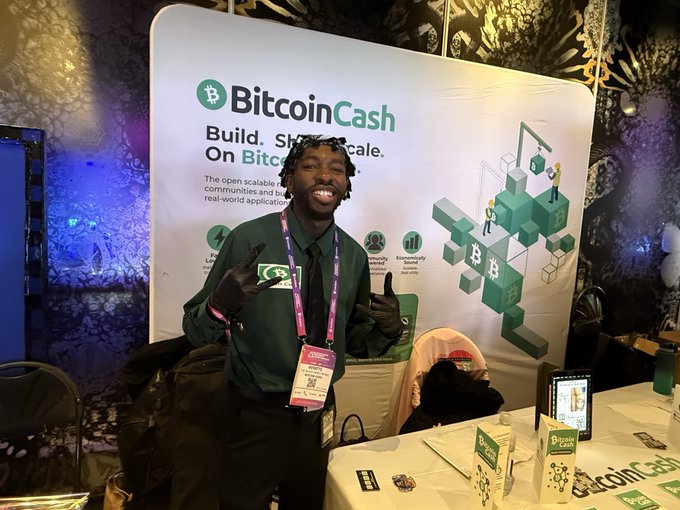 A man in a dark shirt and tie stands smiling in front of a Bitcoin Cash promotional banner that reads 'BitcoinCash - Build, S[cale]. On Bitc[oin Cash]' at what appears to be a crypto conference booth.