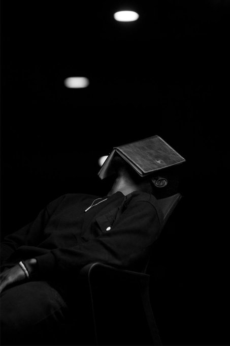 A moody black-and-white photograph of a person sitting in a chair wearing a book or folder over their face, with lit candles and overhead lights in the dark background.