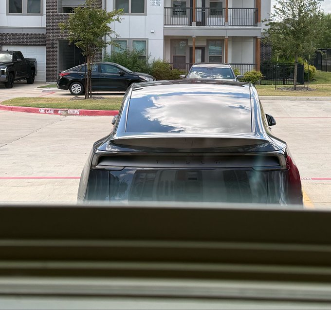 A view from a window looking at a black Tesla Model 3 parked in an apartment complex parking lot.