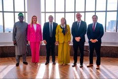A formal group photo of six people in business attire standing together in a modern room with large windows.