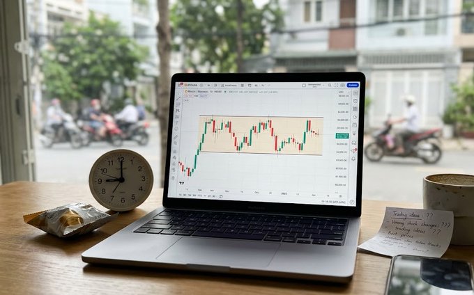 A MacBook laptop on a wooden desk displaying a cryptocurrency trading chart on TradingView, accompanied by a vintage analog clock, a snack wrapper, a coffee mug, and handwritten notes about trading ideas, with a street scene visible through the window behind.