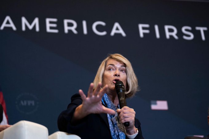 A blonde woman speaks into a microphone at a podium with the words 'AMERICA FIRST' displayed in the background.