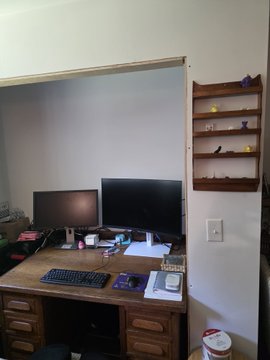 A wooden desk setup featuring dual monitors, a keyboard, and various items, with a decorative bird shelf mounted on the adjacent wall.