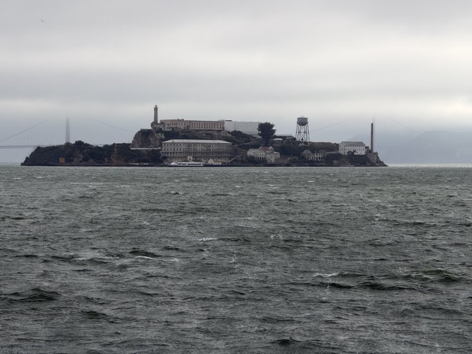 The image shows a large body of water with an island in the middle, surrounded by clouds. The island appears to be made of concrete or stone, possibly resembling a castle-like structure. In the background, there are buildings visible on the island, which adds to its architectural appeal. The post suggests that if you don't buy some mullet from Sol's market, you might end up at this island.