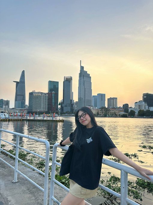 A selfie photo of a young woman wearing glasses and a black t-shirt posing against a waterfront railing with the Ho Chi Minh City skyline, including the Bitexco Financial Tower, visible at sunset in the background.