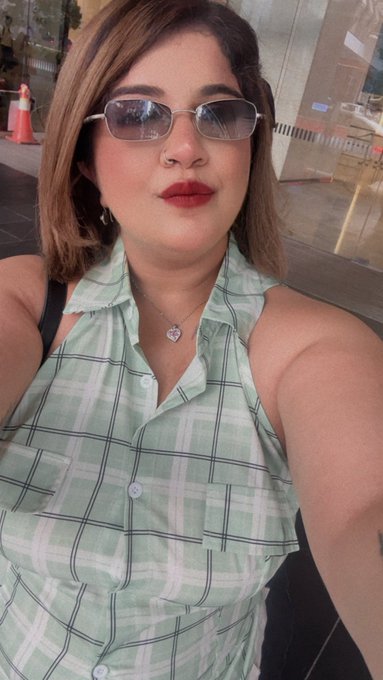 A selfie of a woman with shoulder-length brown hair wearing rectangular sunglasses, red lipstick, and a green and white plaid sleeveless button-up top, with a pendant necklace.