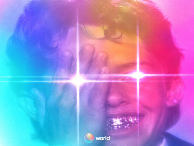 Close-up of a person's face with a hand raised, bathed in neon blue, pink, and purple light with bright lens flares, branded with 'world' and a colorful logo.