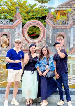 A group of four young people poses together for a photo outdoors, standing in front of an ornate stone fountain with a circular window and traditional architecture visible in the background.