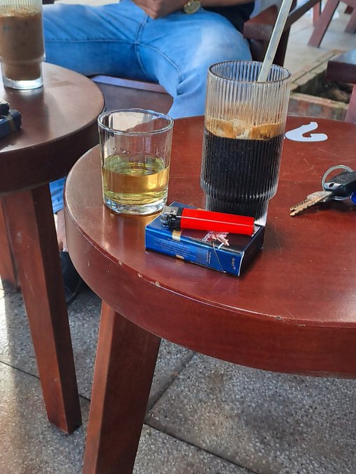 A wooden cafe table holds a glass of dark coffee, a small glass of yellow liquid, a red lighter, keys, and a pack of cigarettes, with a person in blue jeans seated behind.