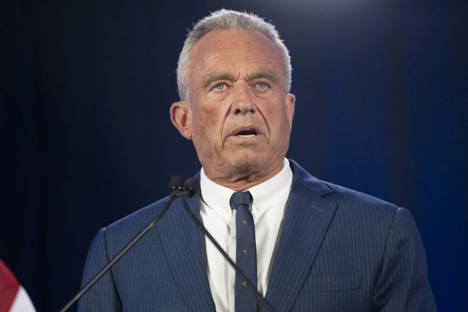The image features a man wearing a suit, standing at a podium with his hands behind him as he speaks into a microphone. The background of the image is blue, which contrasts with the man's dark suit. There are no other objects or people in the image besides the speaker and the microphone. The text "RFK JR: I PUT MOST OF MY WEALTH INTO BITCOIN. I AM FULLY COMMITTED." appears to be related to the speaker, possibly indicating that he is a prominent figure in the Bitcoin community or has made significant contributions to the cryptocurrency space.