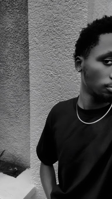 A black and white portrait of a man wearing a black t-shirt and a silver chain necklace standing against a textured wall.