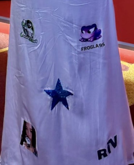 A wrinkled light purple dress is displayed with various crypto project logos taped or printed on it, including "FROGLABS" with a purple frog mascot, a green frog character, a blue star, a partially visible image, and "RN" lettering. The image illustrates the caption's point of poorly executed crypto marketing—an unconventional dress ad where sponsor logos are awkwardly placed and difficult to identify, representing a wasteful and ineffective use of advertising spend.