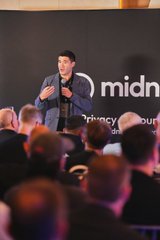A man speaking into a microphone on a stage with a 'midnight' branded backdrop, addressing an audience.