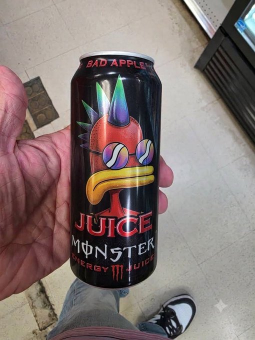 A hand holds a black can of 'BAD APPLE JUICE' branded as a Monster Energy Juice drink, featuring a punk-style apple character with a mohawk and sunglasses.