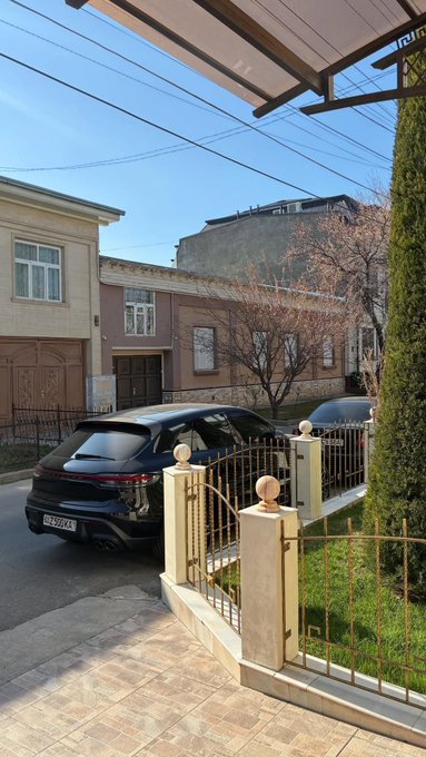 The image shows a residential street scene featuring a dark Porsche Macan (license plate B 2956 KA) parked outside a home with a decorative metal gate and traditional architecture. The scene appears to be a casual "good morning" greeting from the poster, showcasing their car and home in daylight.