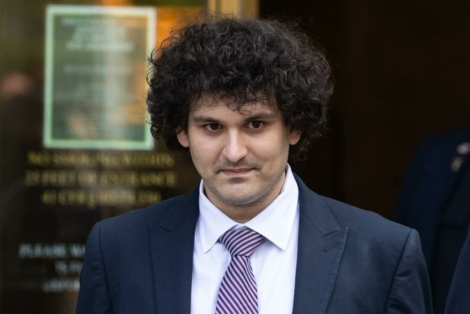 A man with curly dark hair wearing a navy blue suit, white shirt, and red-striped tie stands outside a building with a sign referencing the Department of Justice.