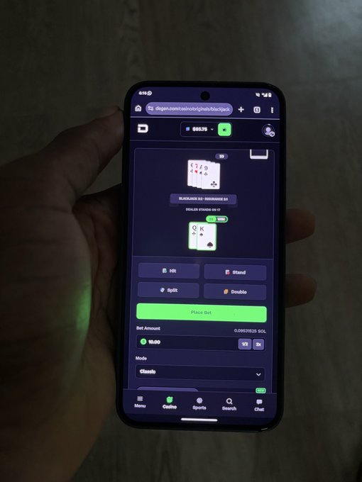 A hand holding a smartphone displaying a crypto blackjack gambling game on degen.com, showing a dealer hand of 6-7-9 and a player hand of Q-K, with options to Hit, Stand, Split, or Double and a $10.00 bet amount in SOL.