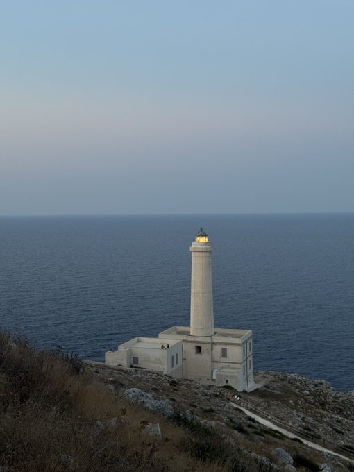 A white lighthouse stands on a rocky cliff overlooking a calm blue ocean at dusk, with a soft gradient sky above.