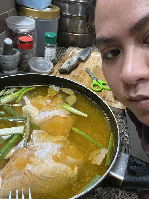 A young woman poses beside a pan of simmering chicken broth infused with green onions, bay leaves, and ginger, while spice jars (including one labeled "5") and green-handled scissors sit on a cutting board behind her—reflecting her prep of a homemade chicken meal to start the day.