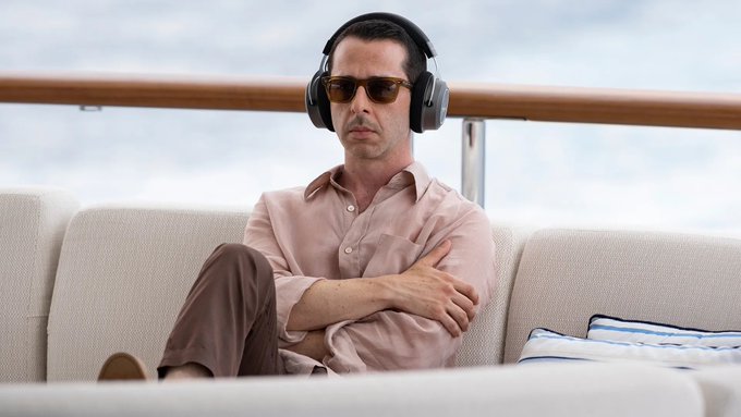 A man wearing large over-ear headphones and aviator sunglasses sits on a cream-colored outdoor sofa on what appears to be a yacht deck, with his arms crossed.