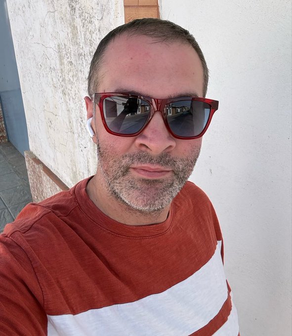 A selfie of a middle-aged man with a short beard, wearing red-framed sunglasses, white wireless earbuds, and a red and white striped t-shirt, standing outdoors against a textured white wall.
