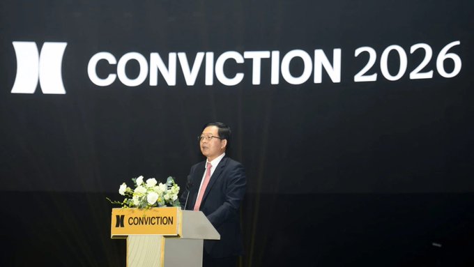 A man in a dark suit and pink tie speaks at a podium with a floral arrangement, standing in front of a large screen displaying "CONVICTION 2026."