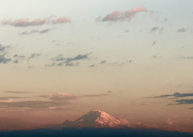 A snow-capped mountain peak rising above a layer of clouds under a soft pastel sky with pink-tinged clouds at dusk or dawn.