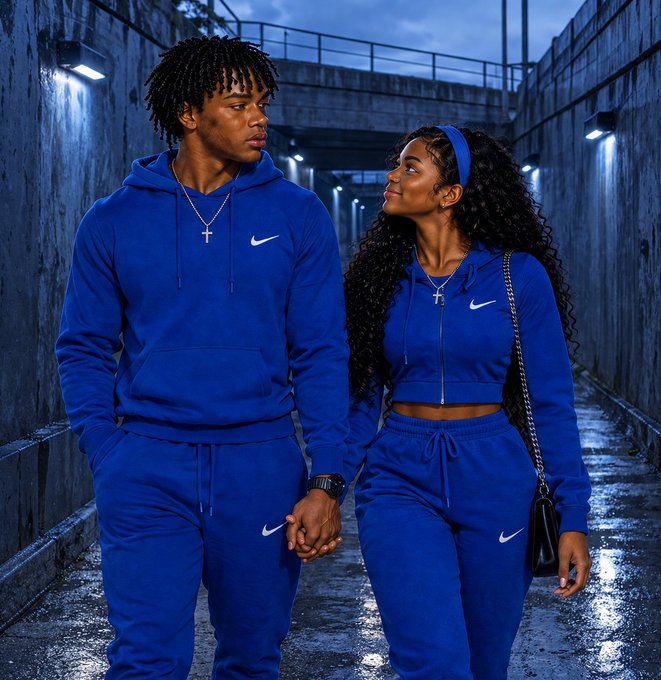 A young couple in matching blue Nike tracksuits holds hands while walking through a dimly lit concrete tunnel, looking at each other affectionately.