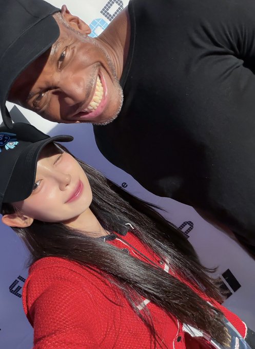 A selfie of a smiling older man in a black cap and a young woman with long dark hair wearing a black cap and a red jacket, posed against a light blue backdrop.