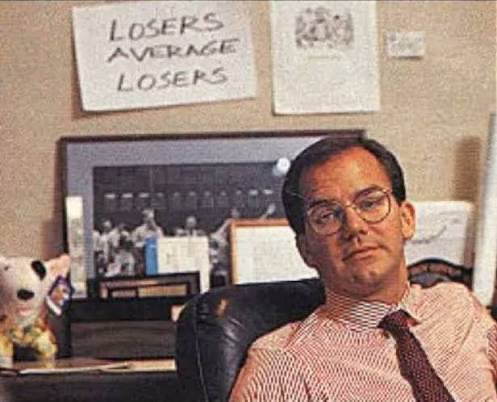 The image features an office setting with a man sitting at a desk wearing glasses and a striped shirt. On the wall behind him are several posters, including one that says "Losses average losers", which seems to be related to the Bitcoin market or cryptocurrency in some way. The man appears to be contemplating something on his computer screen while seated at the desk.