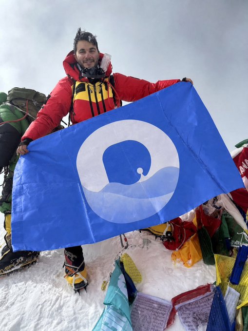 A climber in a red jacket stands on a snowy mountain summit holding a blue flag with a white whale logo, surrounded by colorful prayer flags.