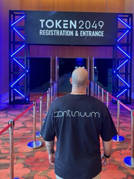 A bald person wearing a black t-shirt with "Continuum" printed on the back walks toward a "TOKEN2049 REGISTRATION & ENTRANCE" sign illuminated by blue neon lights.