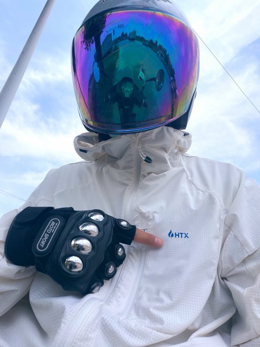 The image features a person wearing a helmet, gloves, and a white jacket with the letters "htx" printed on it. The person appears to be sitting or standing near a pole, possibly at an intersection. The photo also contains text that reads "@HTX_Molly @Ceee333_". This suggests that the image might have been taken by someone named Molly, who is interested in the helmet and gloves worn by the person in the picture.