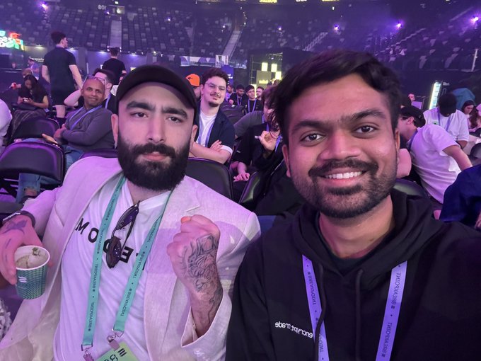 Two smiling attendees pose at Solana's Breakpoint conference, wearing lanyards featuring the "Breakpoint" text, with the man on the right in a black hoodie also displaying "sgornamp" text. The crowded venue with purple-lit ceiling and seated audience reflects the international event atmosphere described in the post about the chaotic journey from India to Abu Dhabi.