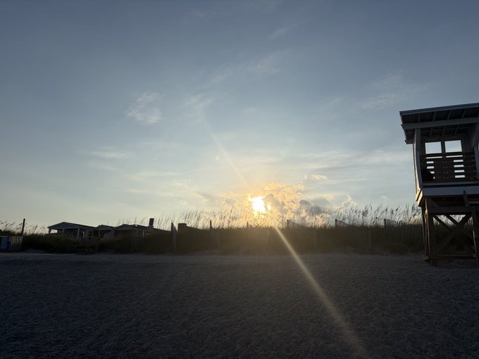 A sunset over a sandy beach, with the sun breaking through a cloud bank, illuminating tall dune grass, while a wooden lifeguard stand and a beach house silhouette the right side of the frame.