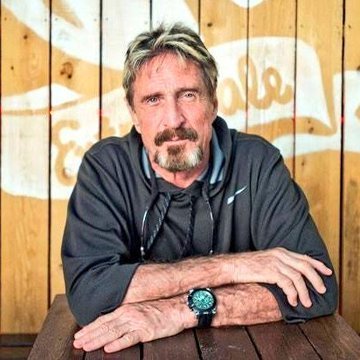 A man with a gray beard and tousled hair, resembling tech entrepreneur John McAfee, sits at a wooden table wearing a dark Nike jacket and a wristwatch, his arms crossed confidently. The reference to his quote about Bitcoin's unstoppable global spread aligns with the image, which visually evokes his well-known, unconventional persona.