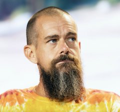 A man with a long brown beard and shaved head wears a bright yellow and red tie-dye shirt, appearing to be Jack Dorsey, the billionaire referenced in the accompanying post about Bitchat, his Bitcoin messaging app being banned in India.