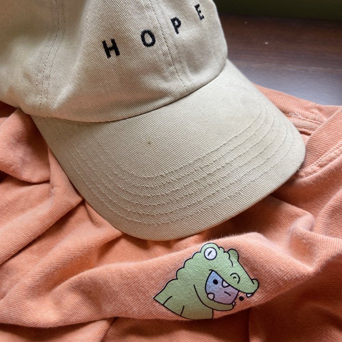 A photo of a beige 'HOPE' cap resting on an orange garment featuring a small green cartoon crocodile character.