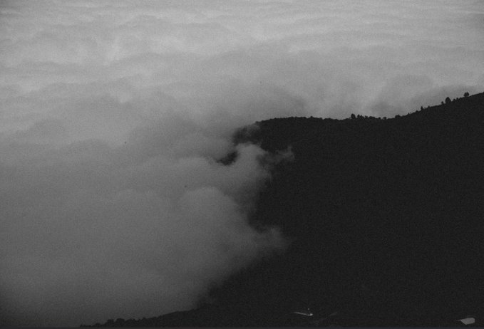 A stark, black-and-white landscape photograph showing a dark mountain ridge or cliff face partially obscured by thick, swirling fog or clouds.