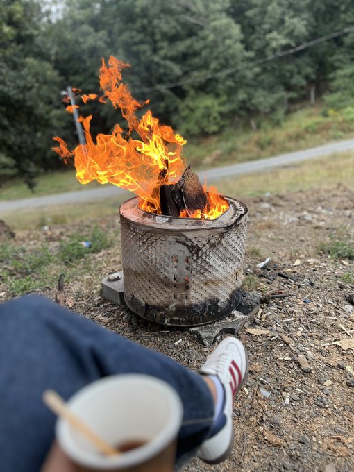 A cozy outdoor scene of a person relaxing by a fire pit with bright orange flames, holding a paper cup, with white sneakers visible against a wooded roadside background.