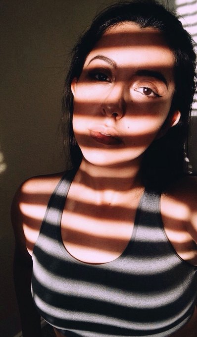 A close-up portrait of a young woman with dark hair wearing a grey striped tank top, with horizontal shadows from window blinds cast across her face and body.