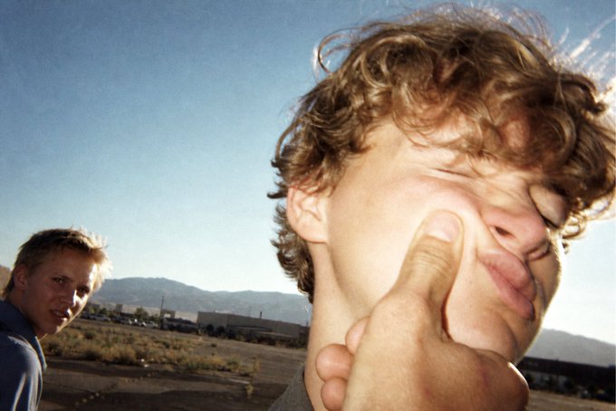 A close-up of a curly-haired person with their eyes closed while a hand covers their mouth/nose, with another blonde person in a blue shirt visible in the background against a desert landscape.