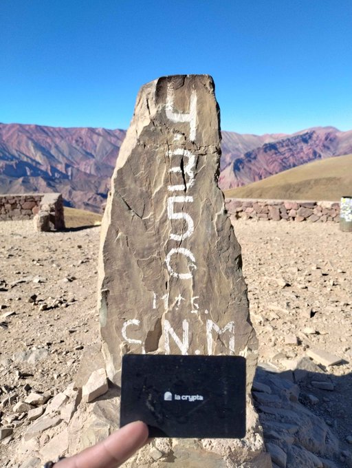 A person holds up a black La Crypta crypto card in front of a tall rock marker painted with '4350 M.S.N.M' (meters above sea level) at a mountain summit with a desert landscape in the background.