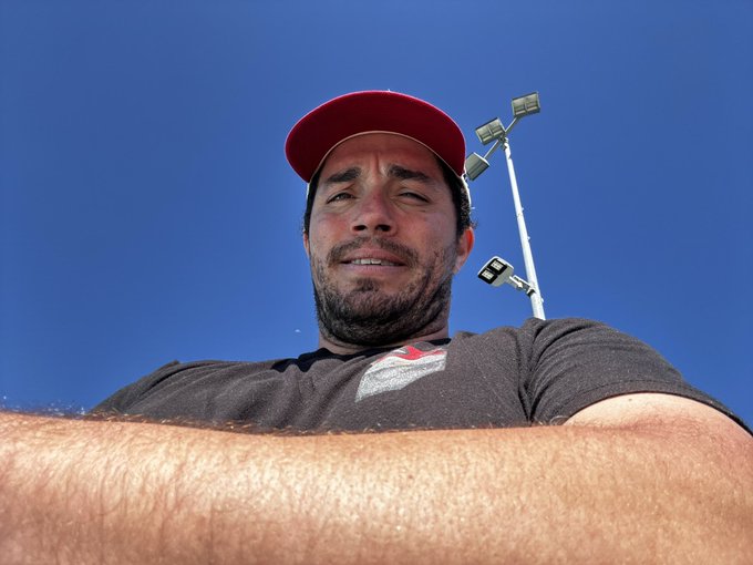 A man in a red cap squints up at the camera against a bright blue sky, with stadium lights visible in the background, giving the low-angle selfie a confident, sun-soaked vibe. The image pairs with the post's bullish call to buy HBAR between .03-.05, matching the relaxed, optimistic energy of someone taking a chance on the dip.