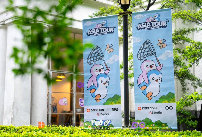 Two light blue banners hang from a lamppost advertising an 'ASIA TOUR' for 'DeepCoin' and '0G', featuring cartoon penguins holding butterfly nets and event logos including 'PUDO EX' and 'AI MEDIA'.